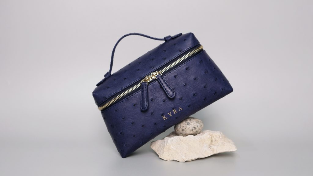 KYRA Picco 19 Navy Ostrich leather bag with distinctive quill follicles and gold hardware accents, showcased against a grey background.
