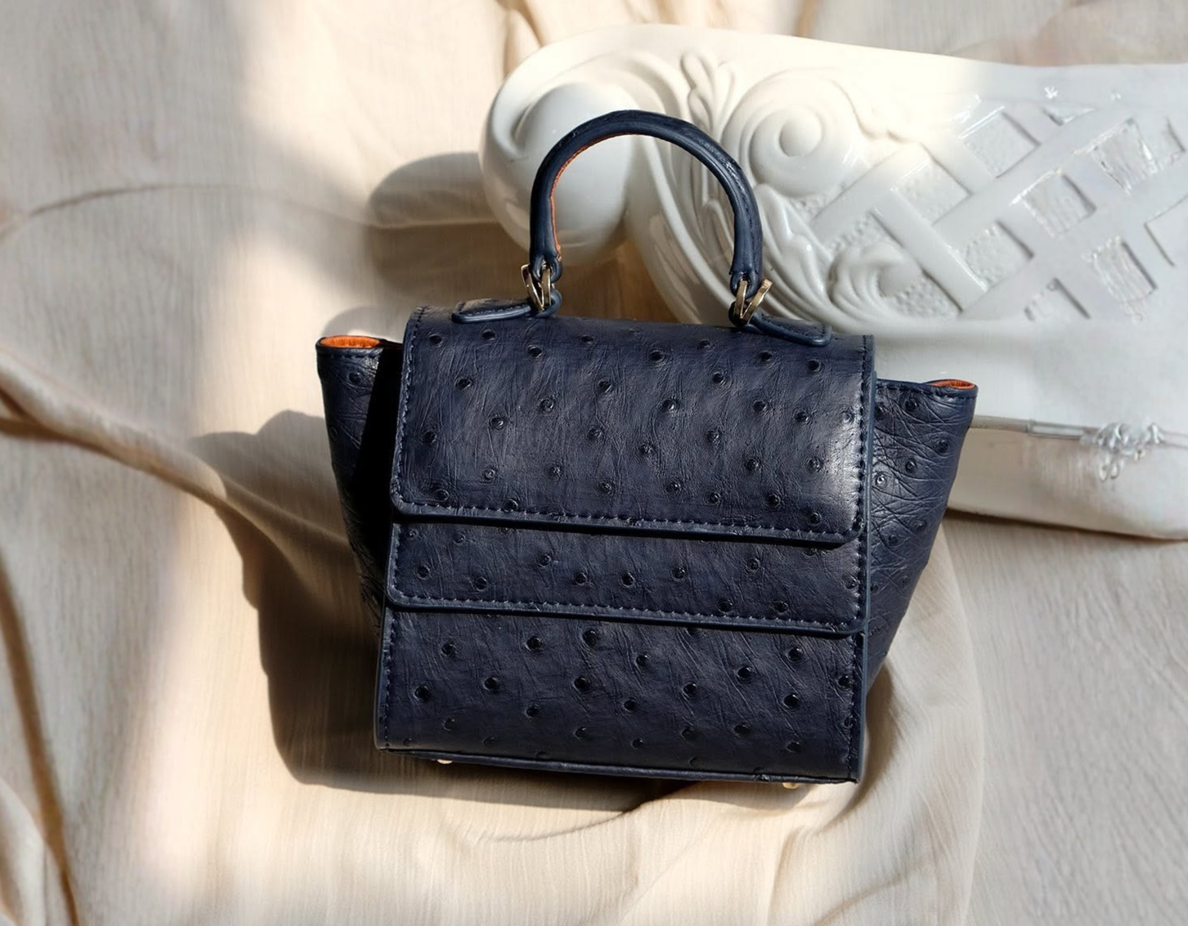 Rubby small navy ostrich leather handle bag with a unique layered closure and structured side wings resting on a soft neutral fabric.