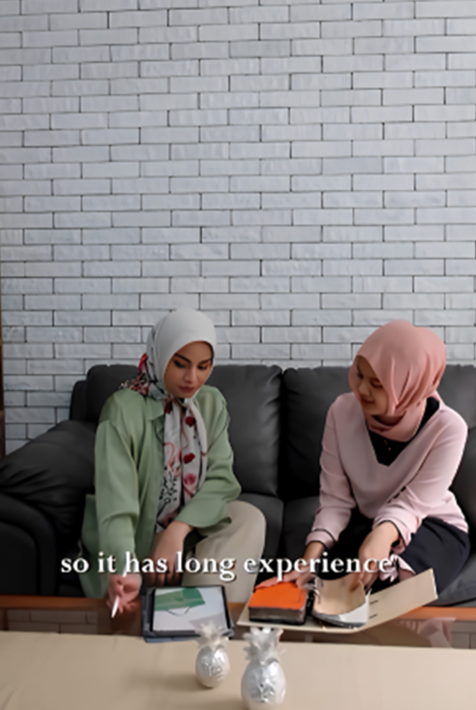Two consultants in hijabs reviewing a leather swatch catalog for Cardina.id, KYRA's specialized custom bag manufacturing service, featuring a legacy of craftsmanship since 1985.