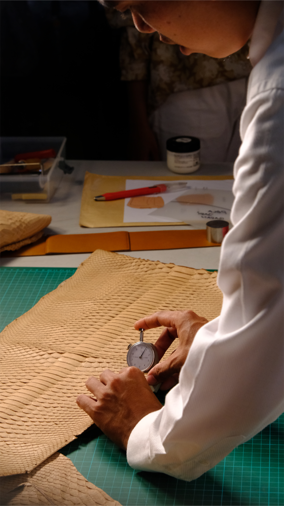 A close-up of a highly skilled KYRA artisan, possessing over a decade of experience, carefully using a precision gauge tool to measure a natural-toned exotic python leather skin. His focus on his task illustrates the meticulous pengerjaan tangan involved in creating a unique personalized luxury item. The scene is set on a traditional workbench with crafting tools.