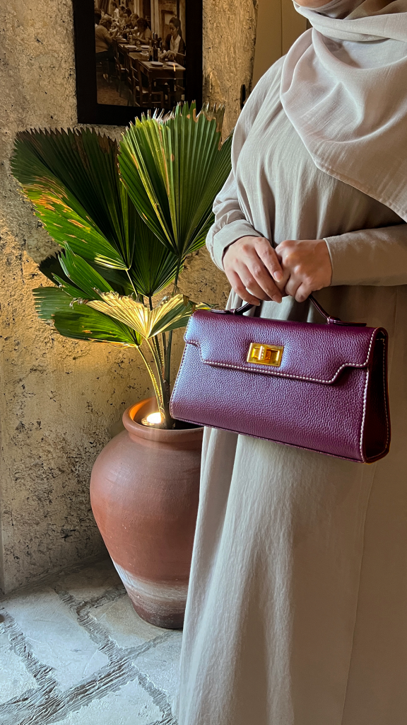 A lifestyle shot of a woman wearing a neutral-toned hijab holding the Luna 25 Burgundy Calf leather handbag by KYRA. The structured bag features a rich burgundy color and elegant gold clasp, showcasing a sophisticated personalized handbag option.