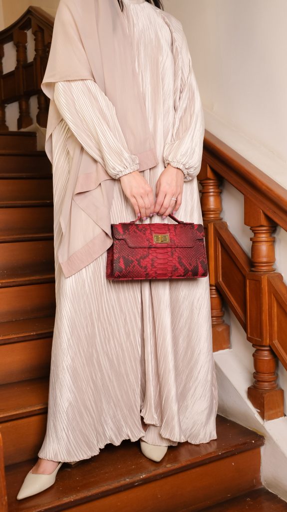 A woman in a beige pleated abaya holding a Burgundy python exotic leather bag called the Luna 29 while standing on a wooden staircase.