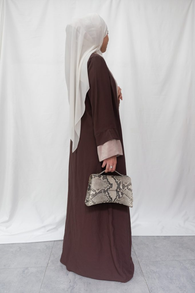 A woman wearing a brown abaya and white hijab holding a natural python exotic leather bag called The Bridge bag with an elegant curved silhouette.