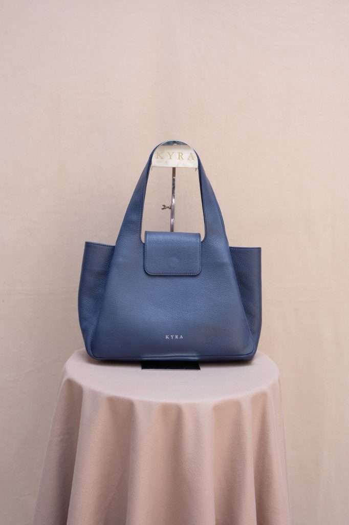 The KYRA Alma premium tote bag in navy blue leather represents our quality work bags featuring a semi structured silhouette and a secure magnetic flap closure displayed on a round table stand.