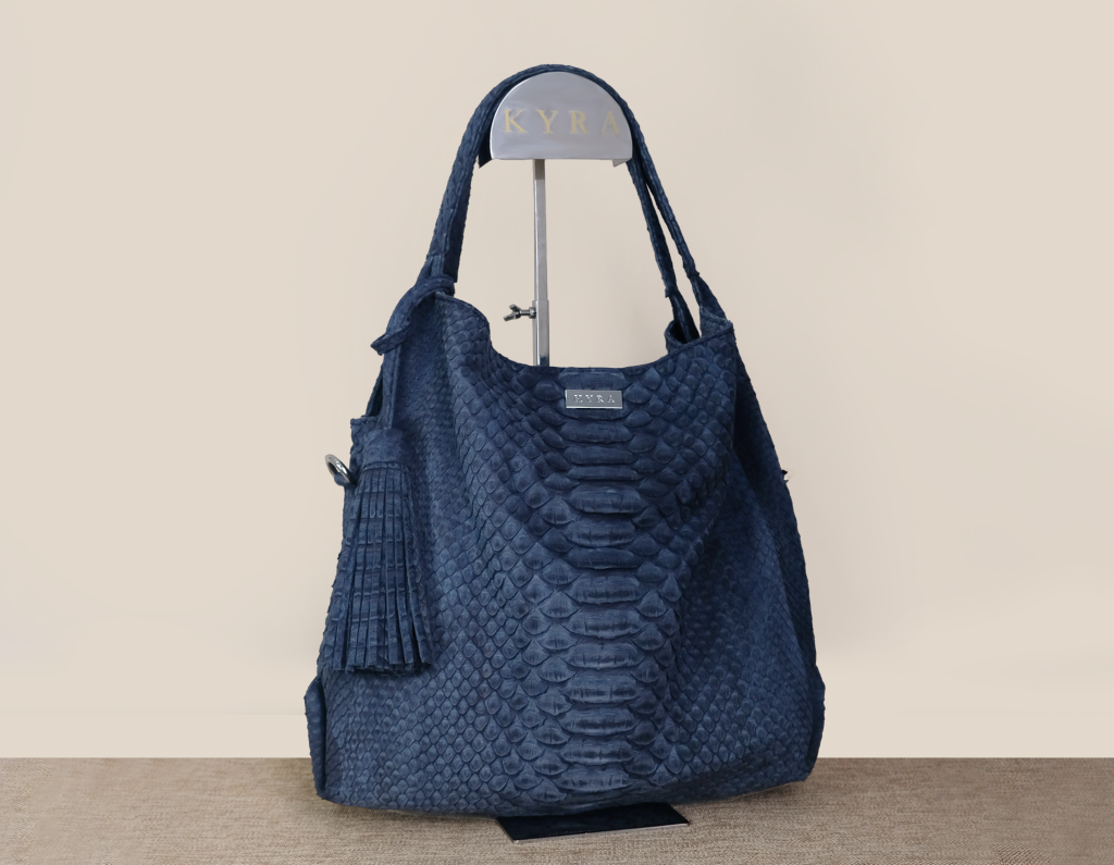 The KYRA Ursula premium tote bag in navy blue exotic python leather represents our quality work bags featuring a soft flexible structure and a matching leather tassel hanging on a display stand.