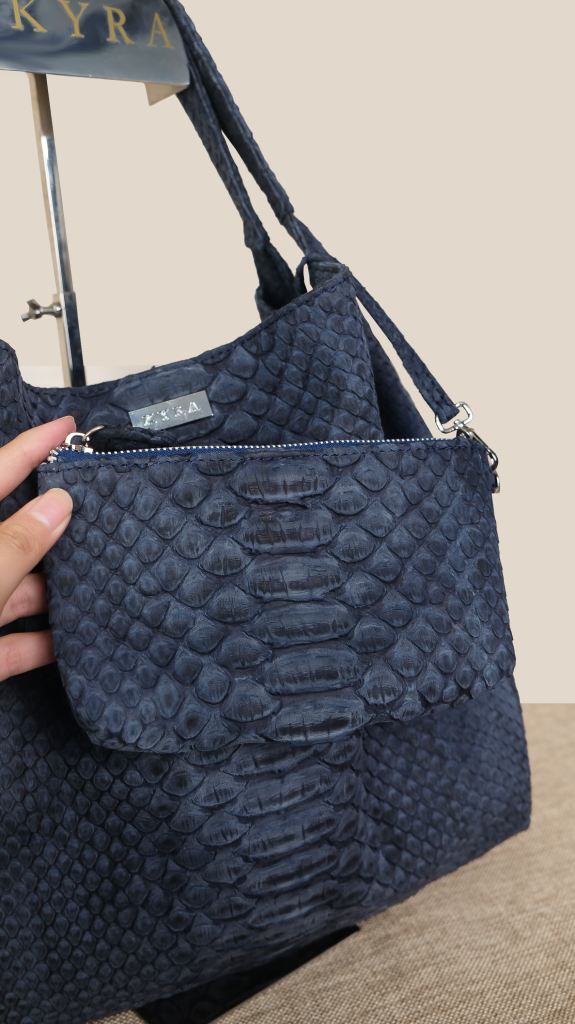 A close up of a hand holding the navy blue removable free matching pouch made from premium exotic python leather attached to the KYRA Ursula tote bag.