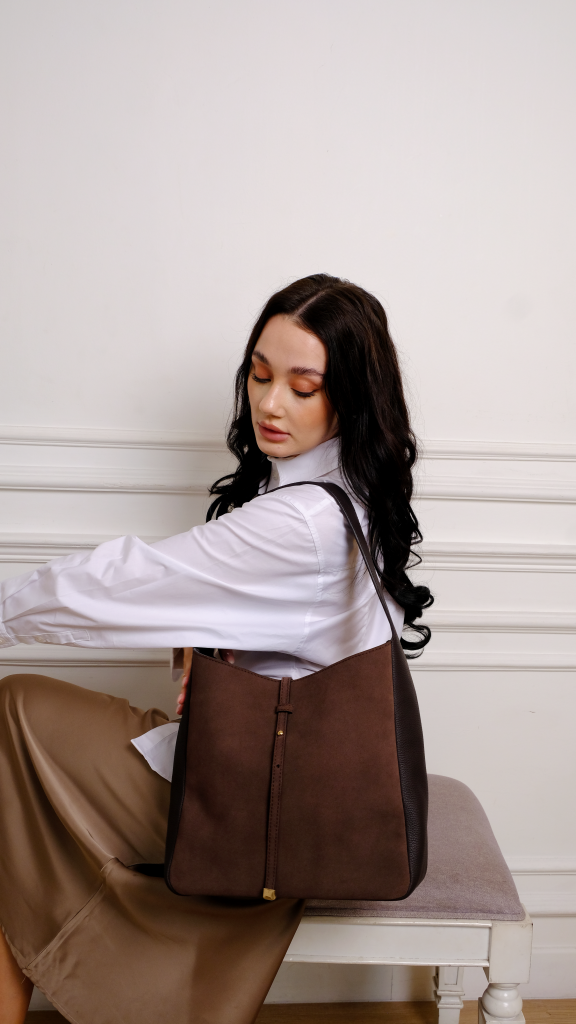 A woman wearing a white button up shirt holding a luxury KYRA Alia Large shoulder bag crafted from dark brown suede and calf leather with a gold stud accent.