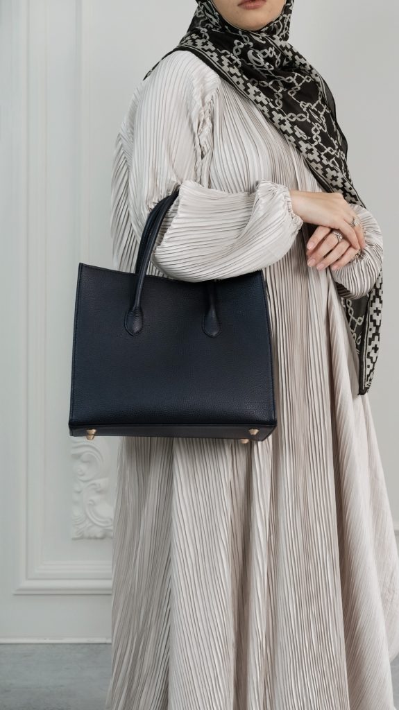 A woman wearing a pleated cream dress and patterned hijab, holding a structured KYRA Roomy tote bag in black pebbled calf leather with gold bag feet.
